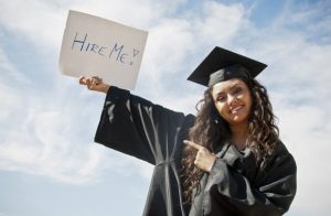 Graduate in cap and gown holding sign that says, "hire me!"